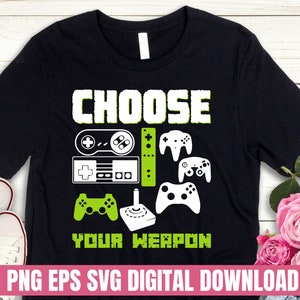Op de afbeelding: Zwart T-shirt met de tekst "CHOOSE YOUR WEAPON" in wit. Het shirt heeft een groene, witte en zwarte videogamecontroller afbeelding.
