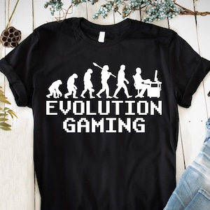 Peut inclure: T-shirt noir avec un graphique blanc de l'évolution de l'homme, se terminant par un homme assis devant un ordinateur. Le texte "EVOLUTION GAMING" est situé sous le graphique dans une police de caractères pixélisée.