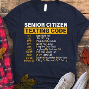 May include: A navy blue t-shirt with a white graphic that says "Senior Citizen Texting Code" and lists common texting abbreviations and their meanings for older adults.