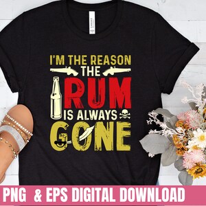 Op de afbeelding: Zwart T-shirt met een gouden en rode afbeelding met de tekst "Ik ben de reden dat de rum altijd op is". De afbeelding bevat ook een fles rum, twee pistolen en een schedel met gekruiste beenderen.