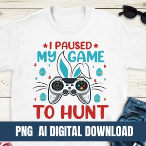 Può includere: Maglietta bianca con una grafica di coniglio blu e bianco e il testo "Ho messo in pausa il mio gioco per cacciare". La grafica include un controller per videogiochi con pulsanti rossi.
