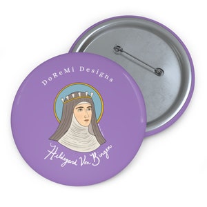 Könnte beinhalten: Ein lilafarbener Knopf mit weißem Rand und dem Text "DoReMi Designs" oben. In der Mitte des Knopfes befindet sich eine Comic-Illustration von Hildegard von Bingen, einer deutschen Benediktinerin, Theologin, Philosophin, Komponistin und Visionärin. Der Text "Hildegard von Bingen" ist in Kursivschrift unter der Illustration geschrieben.