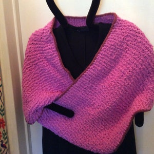 May include: A handmade, pink crocheted shawl with a brown border. The shawl is draped over a black garment on a hanger. The texture of the crochet stitch is visible, creating a cozy and textured appearance.