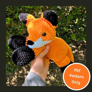 May include: A plush fox toy made of orange, white, and black yarn with a soft, textured finish. The fox features black ears, a white muzzle, and black paws. A circular orange graphic in the corner contains the text PDF Pattern Only.