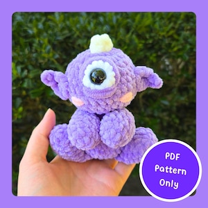 May include: A handmade, plush, purple cyclops creature with a large, black eye and small ears. The crochet monster has a small, light yellow pom-pom on its head and pink cheeks. A circular graphic with the text "PDF Pattern Only" is in the lower right corner.