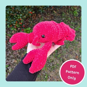 Puede incluir: Un juguete de peluche de langosta rojo brillante con grandes pinzas y un cuerpo texturizado. La langosta tiene ojos negros y se sostiene en una mano. Un círculo rosa con el texto "PDF Pattern Only" está en la esquina inferior derecha.