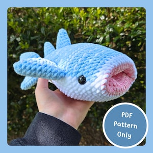 Peut inclure: Un requin-baleine en peluche fait à la main, de couleurs bleu, blanc et rose. Le jouet en crochet doux présente un design détaillé avec des nageoires et une grande bouche. Un graphique circulaire avec le texte "PDF Pattern Only" est dans le coin inférieur droit.
