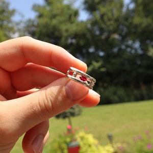 May include: A silver ring with a red gemstone set in the center. The ring is held in a hand.
