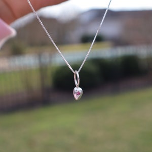 May include: A silver chain necklace with a small heart pendant featuring a pink gemstone in the center.