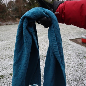 May include: A teal blue knitted scarf is held by a gloved hand. The scarf is folded in half and the ends are hanging down.