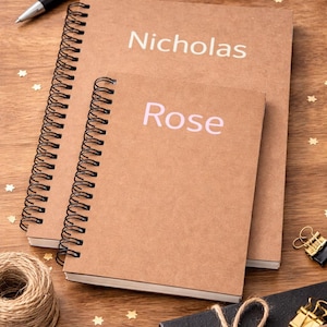 Puede incluir: Dos cuadernos en espiral marrones con los nombres "Nicholas" y "Rose" impresos en las cubiertas. Los cuadernos están sobre una superficie de madera, junto con un bolígrafo, clips y otros artículos de papelería.