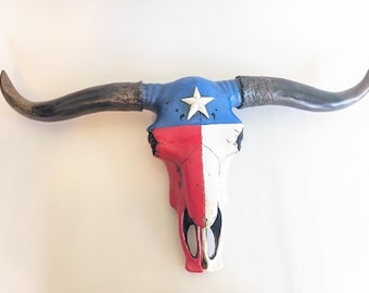 May include: A steer skull wall mount painted with the Texas flag design. The upper portion is blue with a white star, while the lower section is vertically split into red and white panels. The long, curved horns are dark gray with a textured finish.