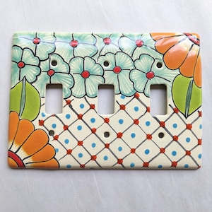 May include: Hand-painted ceramic triple light switch cover. The cover features a floral design with orange and teal flowers, green leaves, and a cream-colored background with a blue and red diamond pattern.