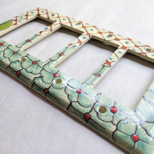 May include: Hand-painted ceramic triple light switch cover. The cover features a floral design in shades of green, orange, and red, with a cream-colored background. The design includes flowers and geometric patterns, adding a decorative touch to any room.