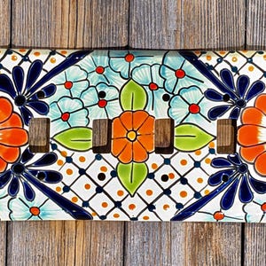 May include: A ceramic light switch cover with a colorful floral design. The design features orange, blue, green, and white flowers on a white background.