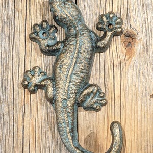 May include: A cast iron gecko hook with a teal and gold finish. The hook is shaped like a gecko with its tail curled into a hook. The gecko is mounted on a wooden surface.