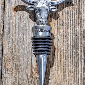 May include: Silver bottle stopper with a Texas Longhorn bull head design. The stopper has a black rubber seal and the word "TEXAS" is engraved on the base.