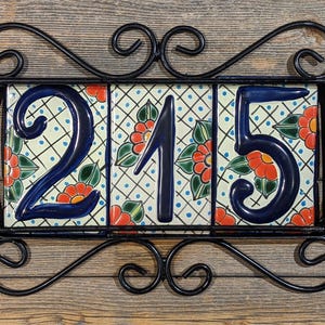 May include: Decorative address plaque featuring the numbers "215" in dark blue, set against a floral tile background. The tiles are framed by a black wrought iron scroll design, mounted on a wooden surface.