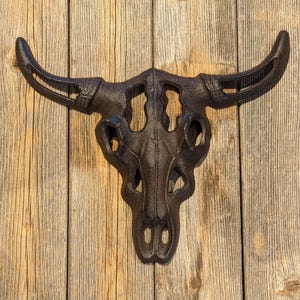 May include: A black cast iron bull skull wall decor with intricate details. The skull is mounted on a wooden surface.
