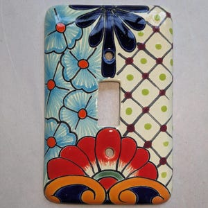 May include: Decorative ceramic light switch cover with a floral design. The cover features a vibrant mix of colors, including blue, red, orange, and green. The design includes flowers and geometric patterns, adding a touch of artistry to any room.
