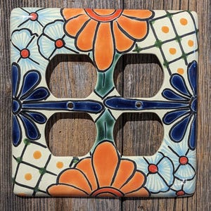 May include: A ceramic light switch cover with a colorful floral design. The design features blue, orange, green, and white flowers on a cream background.