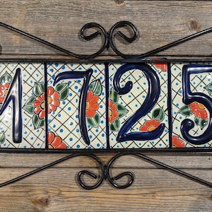 May include: A black metal frame with four ceramic tiles displaying the numbers 1725. Each tile has a white background with blue and green floral designs and red flowers.