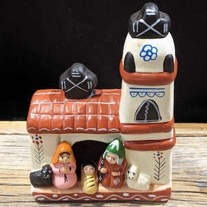 May include: A ceramic nativity scene with a church, three figures, and a sheep. The church is white with a red roof and a black bell tower. The figures are painted in bright colors and are standing in front of the church. The sheep is white and is standing next to the figures.
