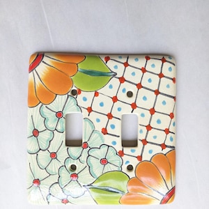 May include: A ceramic double light switch cover with a floral design. The cover features orange, green, and blue flowers, with a cream-colored background and a red and blue grid pattern. The cover has two rectangular openings for the light switches.