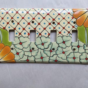 May include: A decorative ceramic light switch plate featuring a floral design. The plate has four switch openings and is painted with a cream background, blue, red, and orange floral accents. The design includes a grid pattern and stylized flowers.
