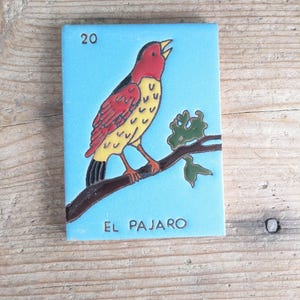 May include: A rectangular, blue tile featuring a colorful bird perched on a branch. The bird has red, yellow, and black plumage. The tile is labeled with the number "20" and the text "EL PAJARO".