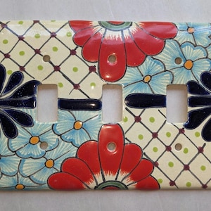 May include: A ceramic light switch plate with a floral design. The plate features red, blue, and navy flowers on a cream background with a diamond pattern. The plate has three switch openings.