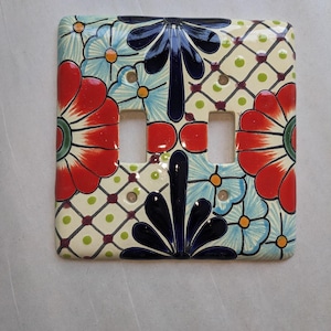 May include: A decorative ceramic light switch cover with a floral design. The cover features red, blue, and black flowers on a cream background. The design includes a grid pattern with green dots.