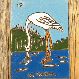 May include: A blue and white ceramic tile with a white heron standing in blue water. The heron is looking down at a red fish in the water. The tile has the number 19 and the words "LA GARZA" printed on it.