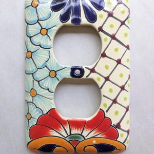 May include: A decorative ceramic double outlet cover featuring hand-painted floral designs. The cover has a cream-colored base with blue flowers, a red and orange floral design, and a geometric pattern. The outlet cover is a unique home decor item.