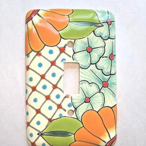 May include: A decorative ceramic light switch cover featuring a floral design. The cover has a rectangular shape with a single switch opening. The design includes orange flowers, green leaves, and blue and red accents on a cream background.