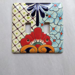 May include: A decorative ceramic double light switch cover. The cover features a colorful floral design with red, blue, and orange accents. The design is hand-painted on a white background with a glossy finish.