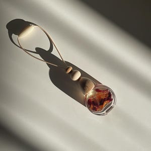 May include: A clear glass car air freshener bottle filled with liquid and dried flower petals. The bottle features a wooden cap, two round wooden beads, and a light-coloured cord for hanging. The design is compact, transparent, and functional.