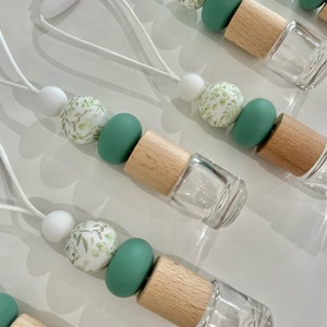 May include: A set of five car air fresheners with a white string for hanging. Each air freshener has a clear glass bottle with a wooden cap, and a string of beads with a white and green floral pattern. The beads are made of silicone and are in white, green, and a light green with a white floral pattern.