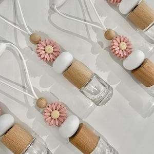 May include: Clear glass essential oil diffuser bottles with wooden caps, white accents, and pink daisy embellishments. Each bottle is attached to a white cord with wooden beads. The bottles are arranged on a white surface.