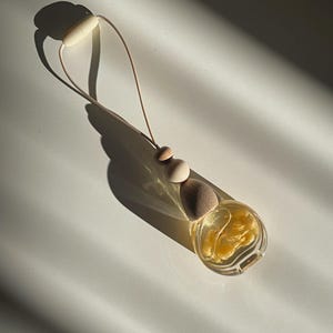 May include: A small, clear glass bottle filled with a pale yellow liquid, featuring a wooden cap and two wooden beads on a light-coloured cord. The bottle is designed for hanging, with a loop at the top and a secure closure mechanism.