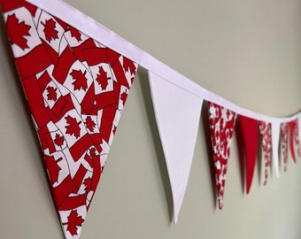 May include: A festive banner featuring alternating red and white pennants. Some pennants display a pattern of red maple leaves on a white background, while others are solid red or white. The banner is strung on a white ribbon, perfect for patriotic celebrations.