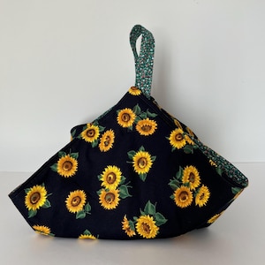 Peut inclure: Un chauffe-bol en tissu avec un fond noir orné d'un motif de tournesols jaunes et de feuilles vertes. L'objet présente une forme triangulaire avec une poignée supérieure pointue faite d'un tissu vert à petit motif floral blanc à l'intérieur.