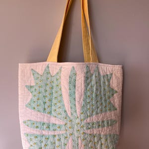 May include: A quilted tote bag with a pale yellow handle and a white body. The body features a large, stylised floral design in light blue with yellow floral accents. The bag is made of soft fabric and has a quilted texture.
