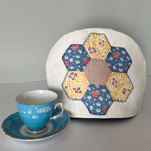 May include: A dome-shaped fabric tea cosy featuring a central hexagon patchwork design in blue, yellow, and white floral patterns, paired with a bright turquoise teacup and matching saucer decorated with white leaf and acorn motifs.