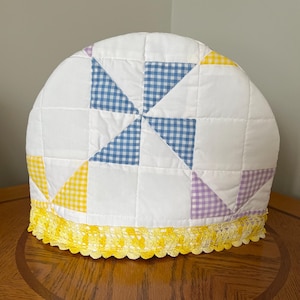 Peut inclure: Un chauffe-thé matelassé blanc avec un motif patchwork composé de triangles en vichy bleu, jaune et violet. Le chauffe-thé est orné d'une bordure en dentelle jaune en bas. Il est posé sur une table en bois.