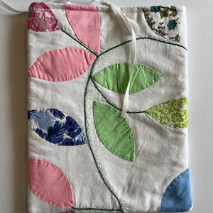 May include: A white fabric pouch with a leaf design. The leaves are in shades of pink, green, and blue, with a green stitched outline. The pouch has a white ribbon closure and is likely handmade.