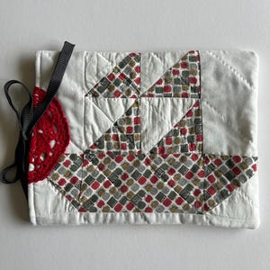 May include: A rectangular quilted fabric pouch featuring a white background with a geometric patchwork design in red, green, and grey. A red crocheted circular accent is attached to the left side, secured by a black ribbon tie closure.