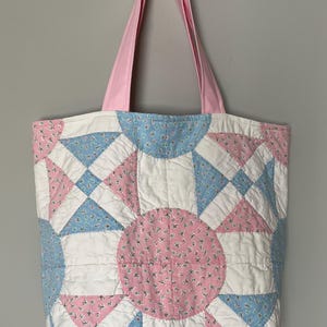 May include: A quilted tote bag featuring a patchwork design of white, light blue, and pink floral fabric. The bag has a central circular motif surrounded by geometric shapes and includes two solid light pink fabric shoulder straps at the top.