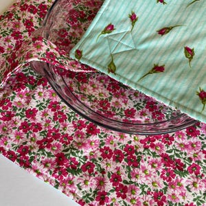 Peut inclure: Un plat de cuisson en verre transparent est placé dans un porte-plat en tissu matelassé. L'extérieur présente un motif floral rose et blanc vif, tandis que les rabats intérieurs sont en tissu rayé turquoise clair avec des boutons de rose.
