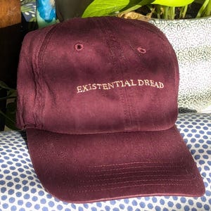 May include: A maroon baseball cap with the text "EXISTENTIAL DREAD" embroidered in white on the front.